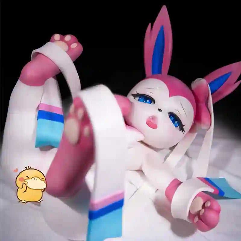 A pink and white anime-style plush doll with bunny ears, blue eyes, and paw-print feet, posed on white bedding against a black background. A small cartoon duck sticker is visible near the lower right area.