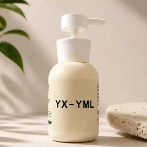 A cream-colored pump lube bottle with black text reading 'YX-YML' on the front, set on a light surface with soft shadows. A green leaf and a textured stone slab are visible in the background.