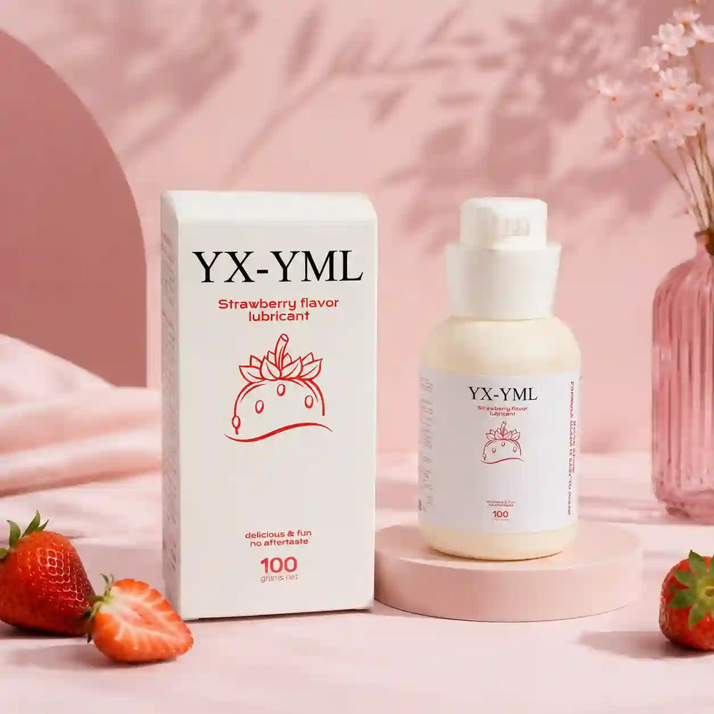 A bottle of YX-YML strawberry flavor lubricant stands next to its white box on a pink surface, with strawberries and a pink floral vase in the background.
